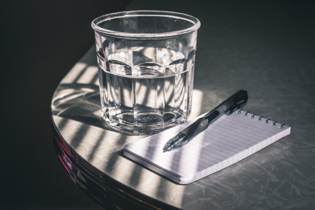 Un verre d'eau à côté d'un calepin et un stylo. Un support pour écrire facile à emporter toujours avec nous pour débuter en rédaction web